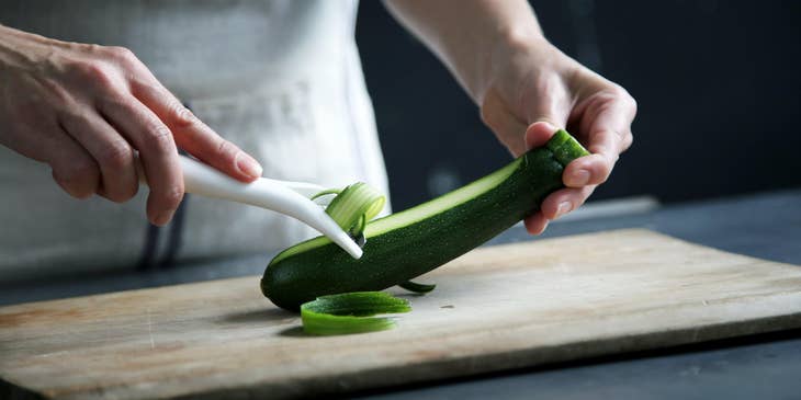 A kitchen staff wearing a white apron peeling a cucumber on a chopping board.