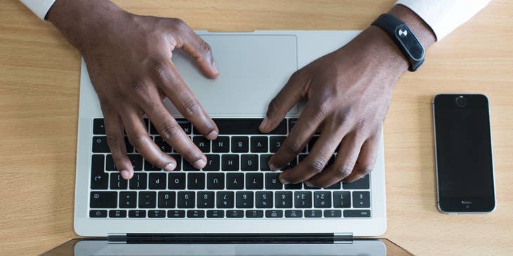Las manos de una persona en una MacBook escribiendo una descripción de empleo con un iPhone al lado.