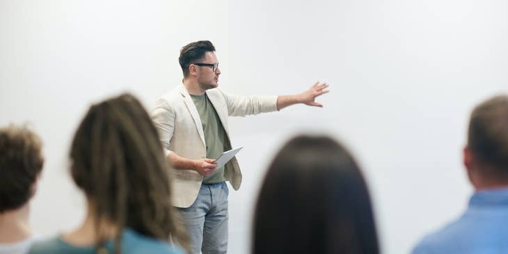 A philosophy teacher holding a tablet and pointing to a wall while students pay attention.