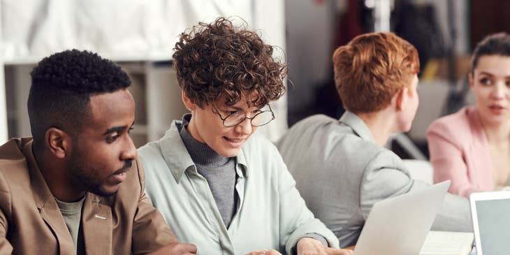 A PMO analyst discussing a project with a co-workers while looking at a laptop.