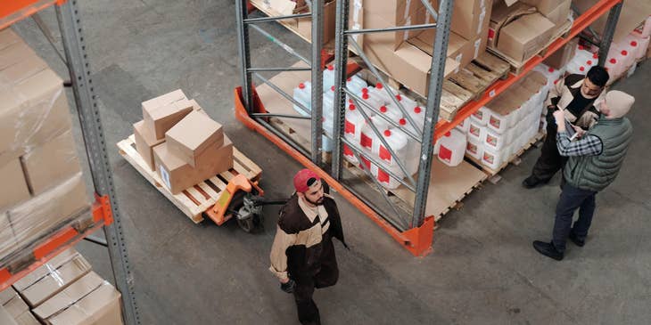 A warehouse sorter moving packages from one spot to another while two co-workers stand nearby discussing work.