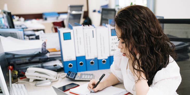 A Female Claims Examiner in an office sitting in front of her laptop and writing on a journal.