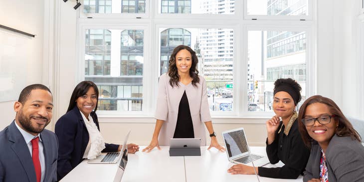 female Customer Service Coordinator smiling and standing while the rest of her team members are sitting inside the meeting room