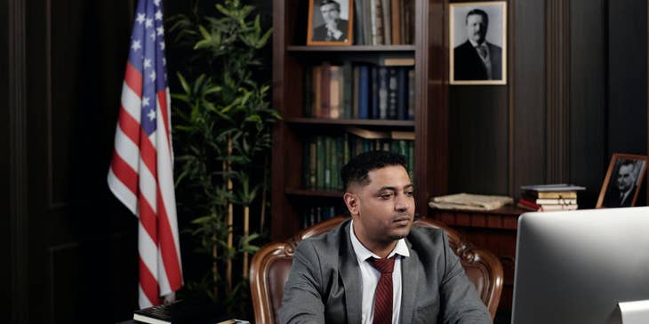 Foreign service officer sitting at his workstation with the bookshelf and the American flag behind him