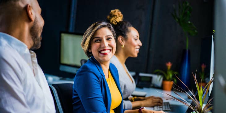female IT Business Analyst sitting at her workstation and sharing a laugh with her colleagues beside her