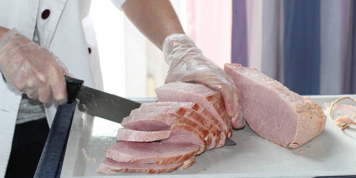 A line cook wearing a white uniform holding a knife and slicing meat on a chopping board.