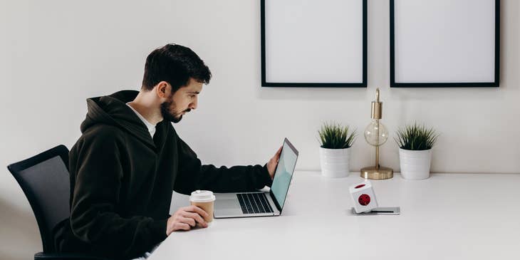 Magento Developer sitting at a white table and looking at his laptop while holding a cup of coffee