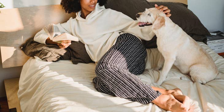 Pet sitter lounging on bed and holding a book while petting a white dog.