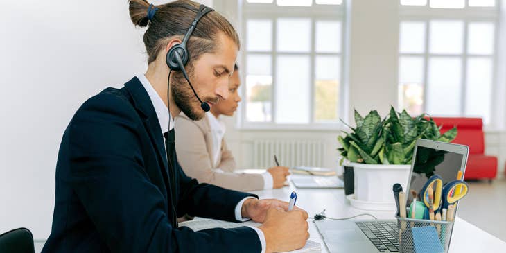Telemarketing executive writing on a piece of paper and wearing a headphone with an open laptop on his desk