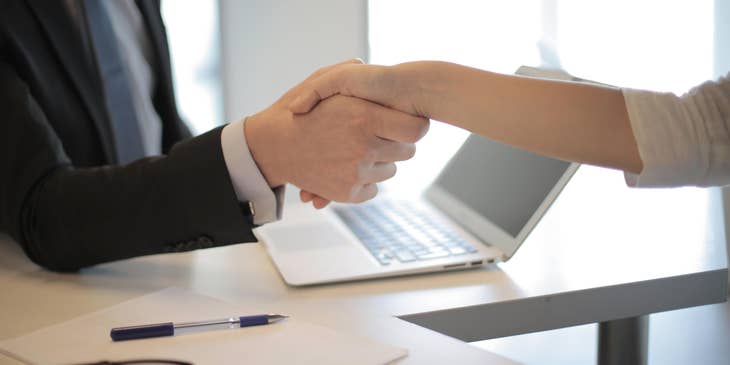 Wealth manager wearing a business suit shaking hands with a client over an open laptop, eyeglasses, piece of white paper and a pen